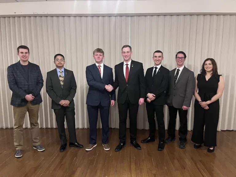 Seven people in formal attire stand in a line as two people in the center shake hands.