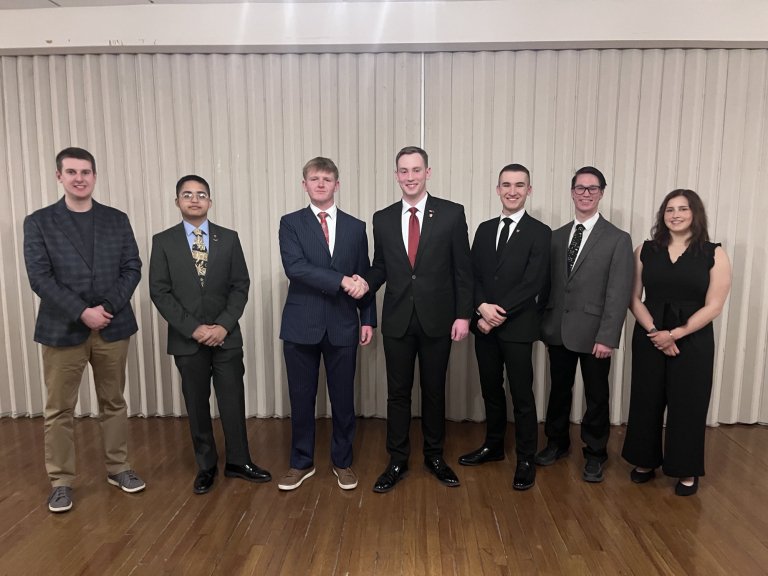 Seven people in formal attire stand in a line as two people in the center shake hands.