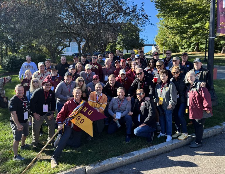 Group of people stand on a lawn holding a Norwich NU 90 flag near trees and a campus walkway.