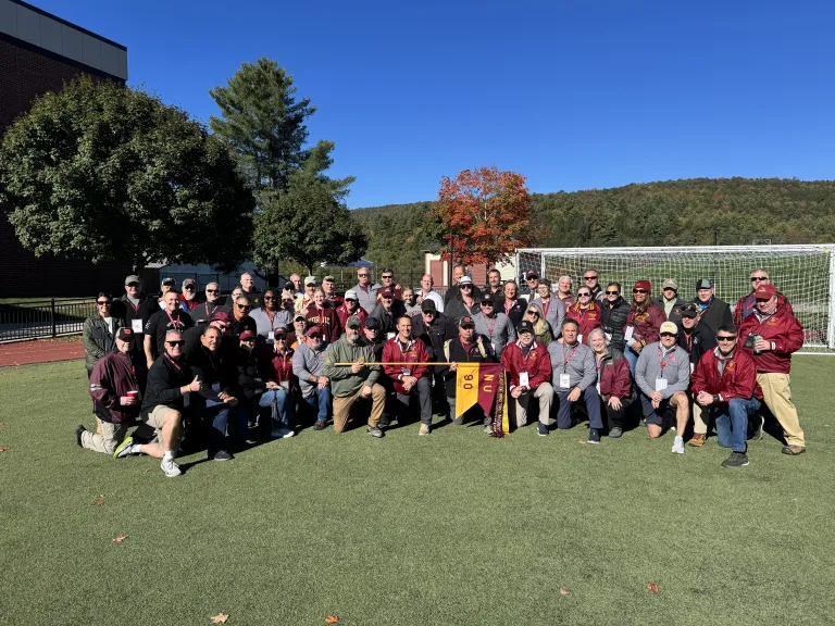 Group of people pose on a turf field holding a Norwich NU 90 banner with a soccer goal and trees behind.