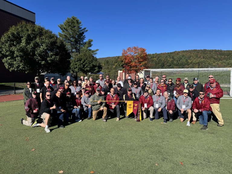 Group of people pose on a turf field holding a Norwich NU 90 banner with a soccer goal and trees behind.