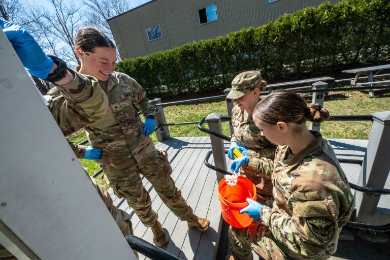 Cadets in camouflage uniforms collect materials in a bucket outside.