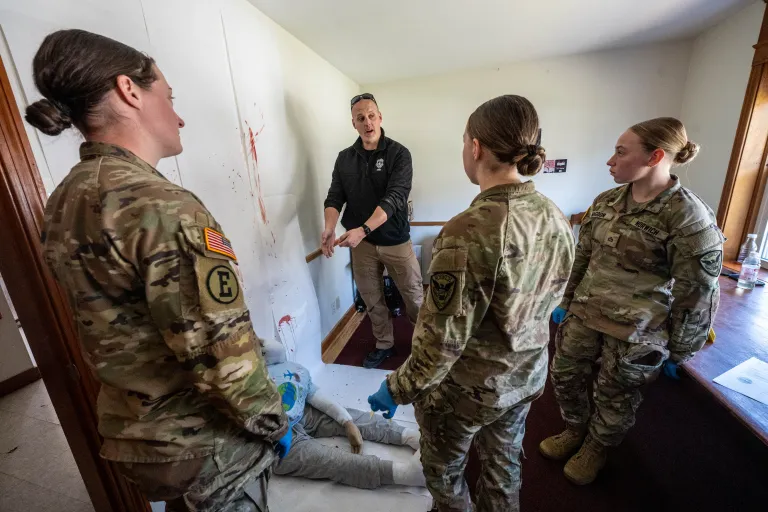 Cadets in camouflage uniforms listen to an instructor beside a training mannequin.