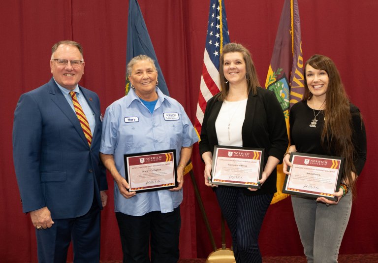 A man stands with three staff award recipients holding Norwich University certificates in front of flags.