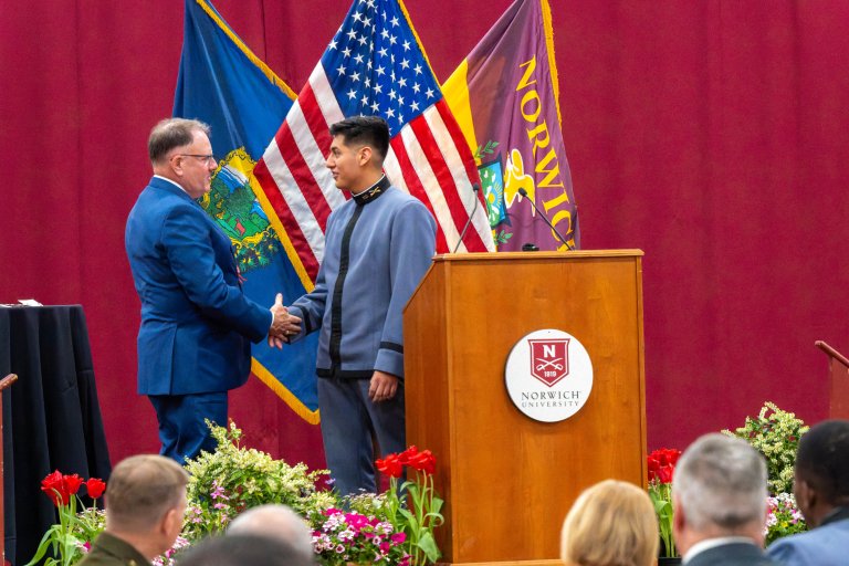 Cadet shakes hands with a man beside a Norwich University podium with American, Vermont, and Norwich flags.