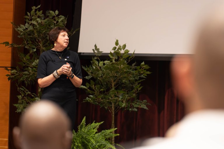 Speaker stands onstage with hands clasped beside a projection screen.