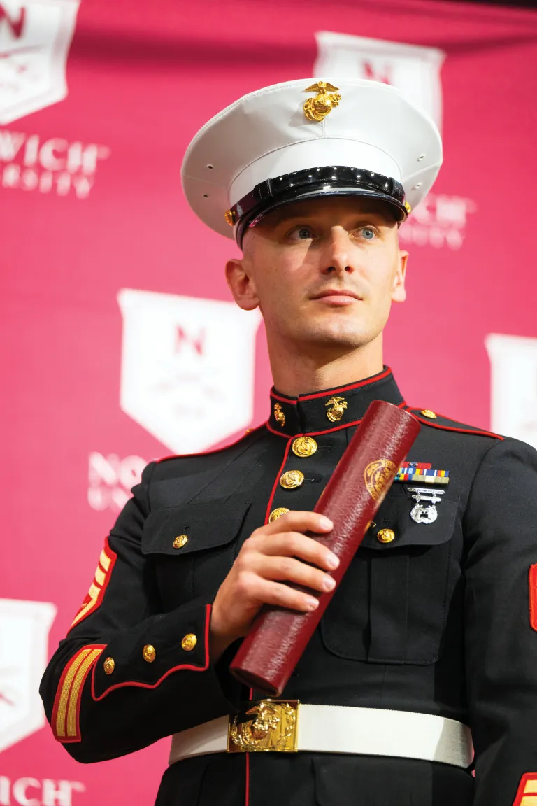 Marine in dress uniform holds a diploma in front of a Norwich backdrop.