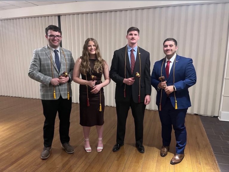 Four students hold gavels and cords while standing in formal attire in a meeting room.