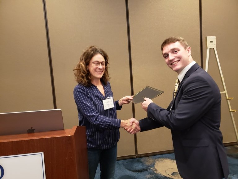 Person receives a certificate while shaking hands beside a podium in a conference room.