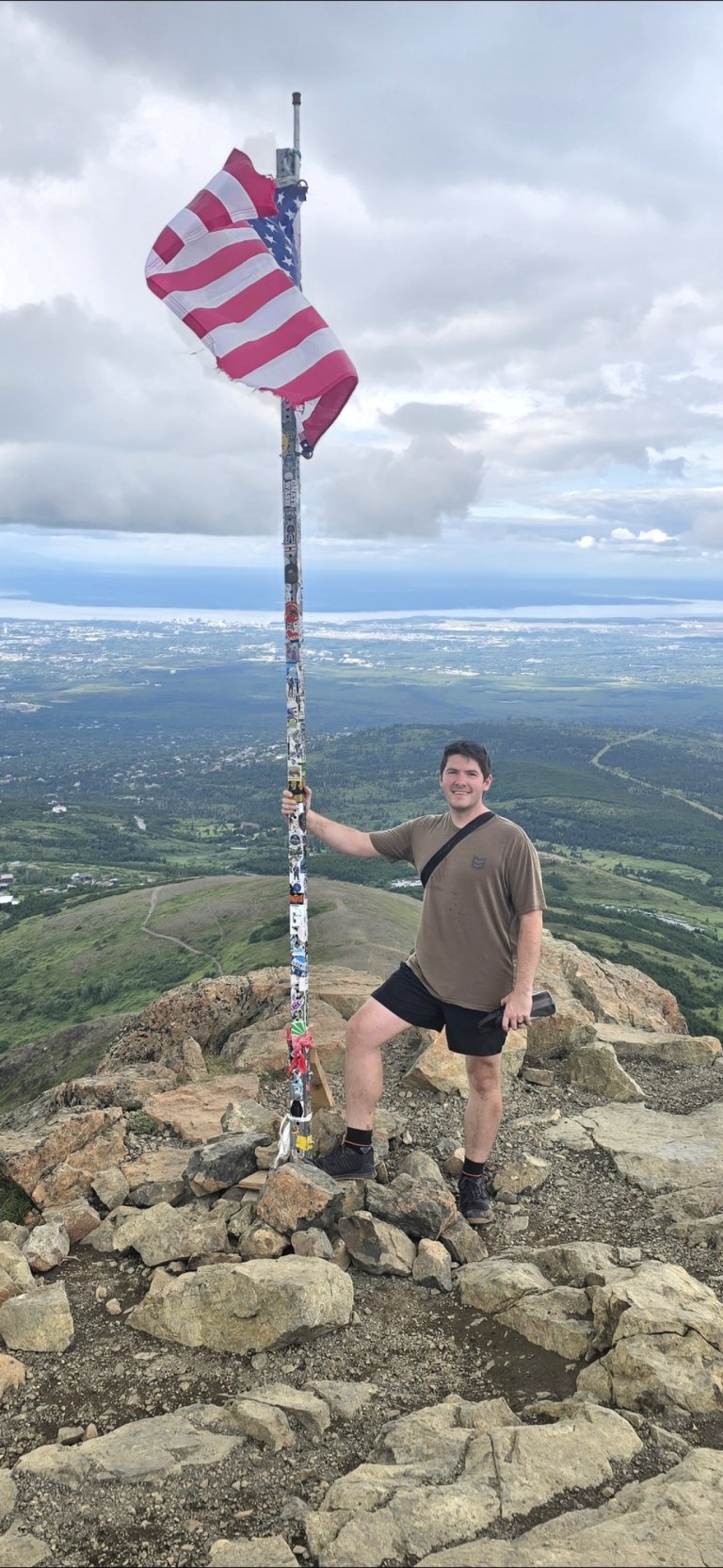 Person stands on rocky summit holding a pole with an American flag above a valley and distant water.