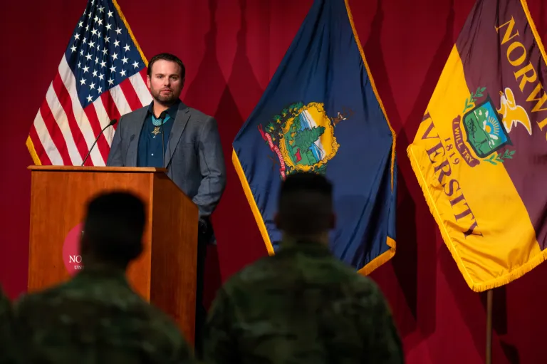 Speaker stands at a Norwich University podium between American, Vermont, and Norwich flags.