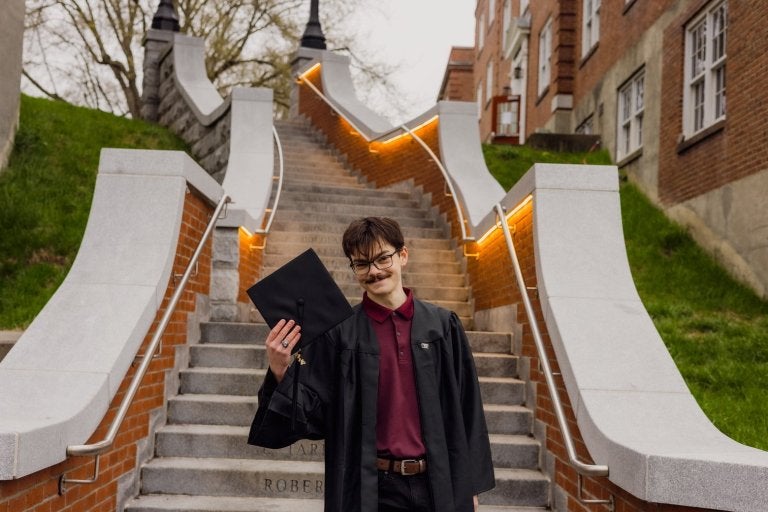 Graduate holds cap while standing on campus stairs between brick buildings.