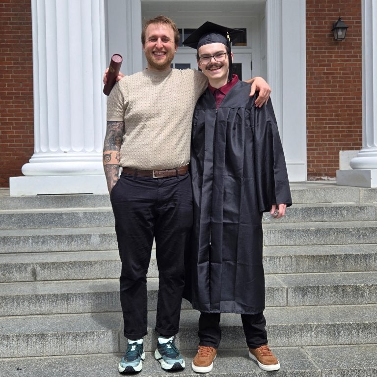 Graduate in cap and gown stands with another person on steps outside a brick building.