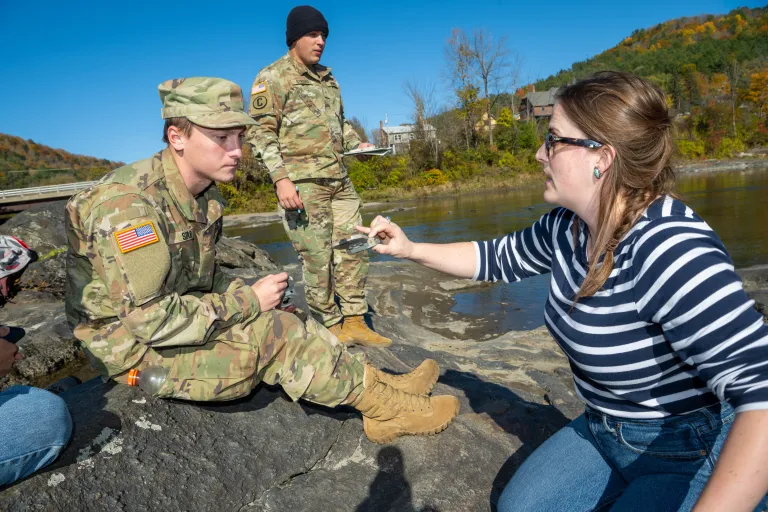 Instructor shows measuring tool to cadets seated on rocks beside a river during outdoor fieldwork.