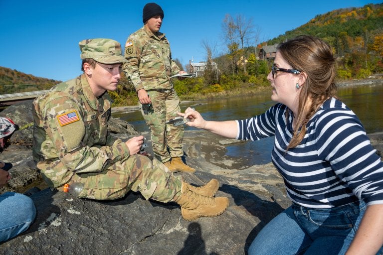 Instructor shows measuring tool to cadets seated on rocks beside a river during outdoor fieldwork.