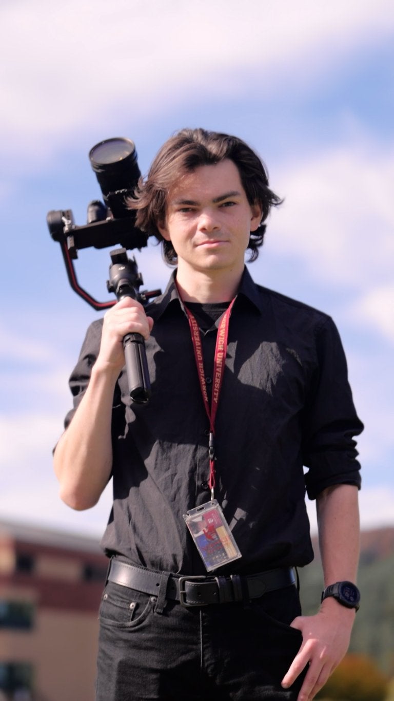 Student holds camera stabilizer outdoors while wearing a Norwich University lanyard.