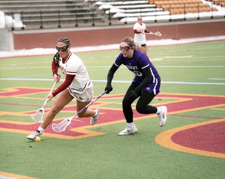 Norwich lacrosse player carries the ball while a Curry defender runs beside them on the field.