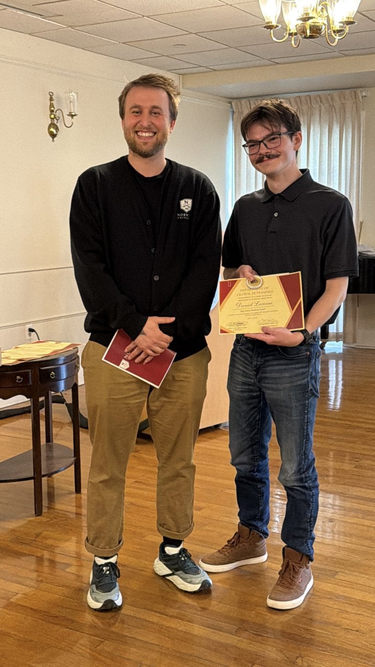 Two people stand indoors while one holds a certificate during an award presentation.