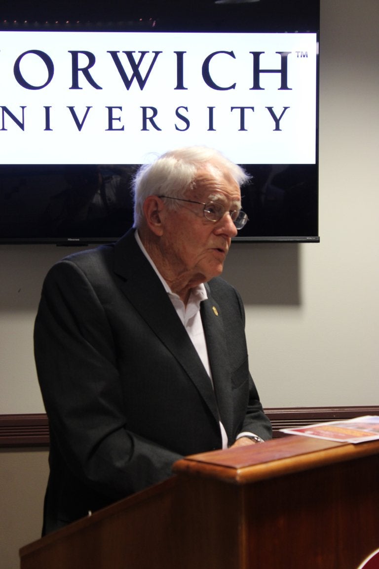 An older man speaks at a podium in front of a screen displaying the Norwich University logo.