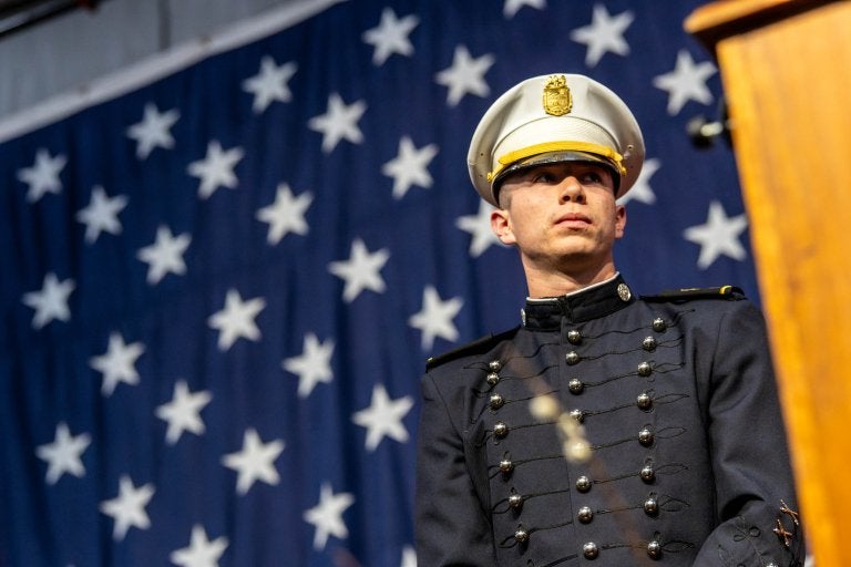 Cadet in dress uniform stands before large American flag during ceremony.