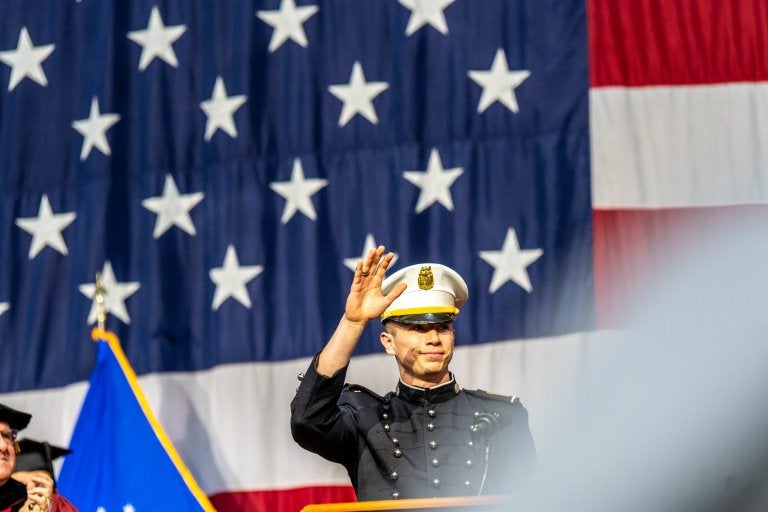 Cadet in dress uniform stands before large American flag during ceremony.