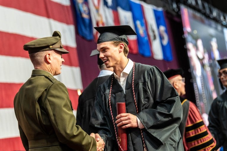 Graduate in cap and gown shakes hands while holding diploma at Norwich University ceremony.