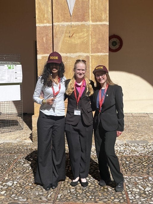 Three students wearing ROOK hats and conference badges stand in front of a stone column outdoors.