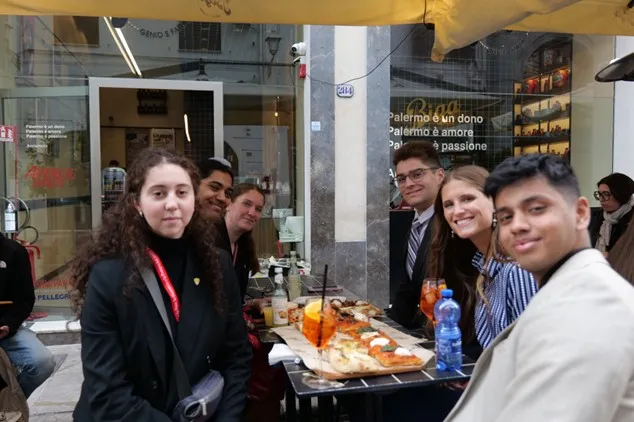Students sit around an outdoor cafe table with pizza and drinks on a city street.