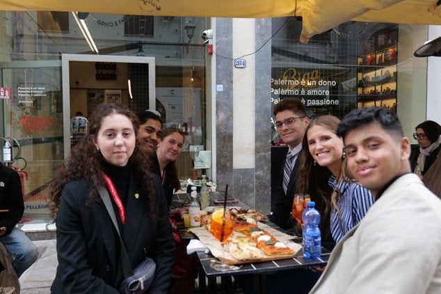 Students sit around an outdoor cafe table with pizza and drinks on a city street.