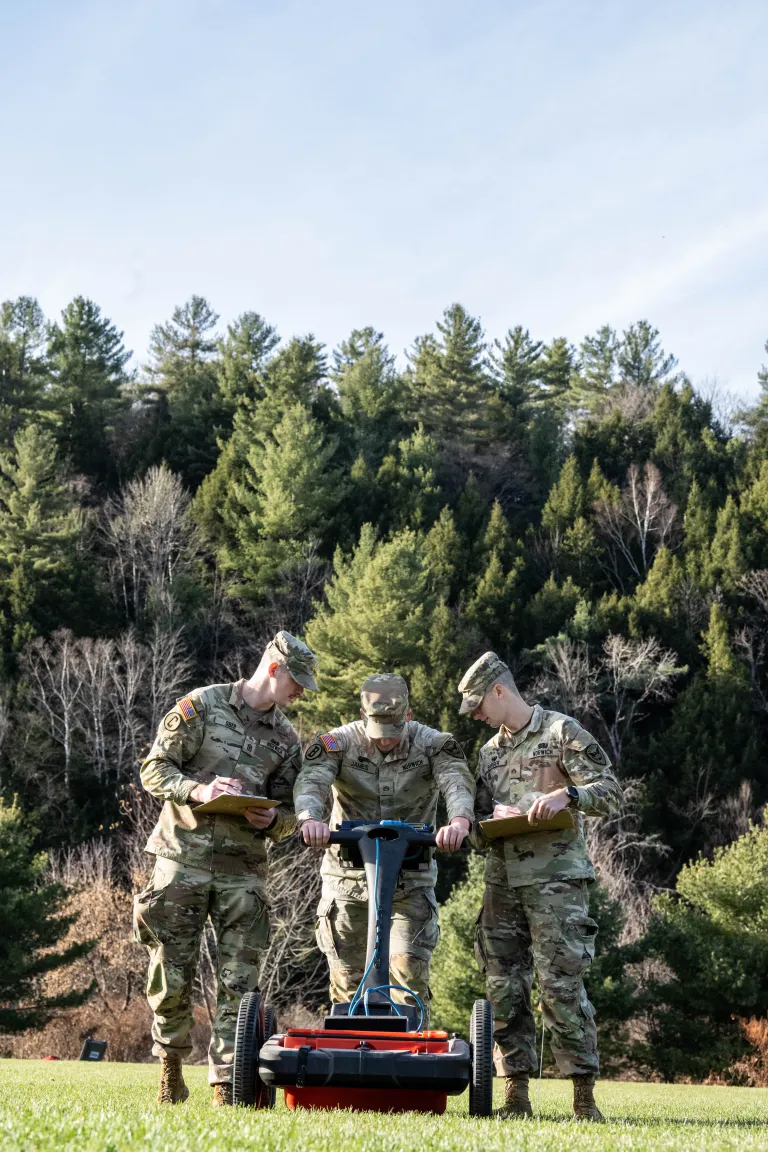 Three cadets use a field surveying device while recording data on a grassy hillside.