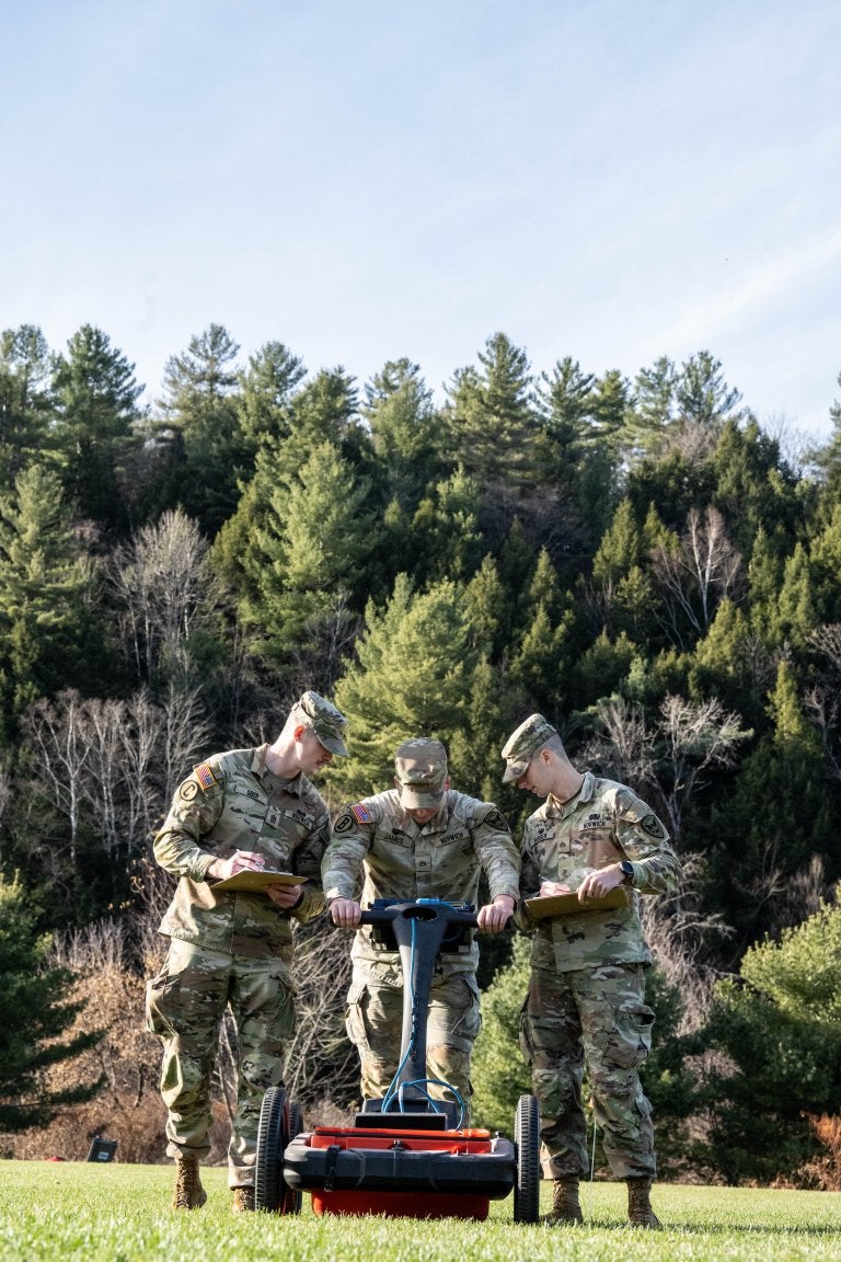 Three cadets use a field surveying device while recording data on a grassy hillside.