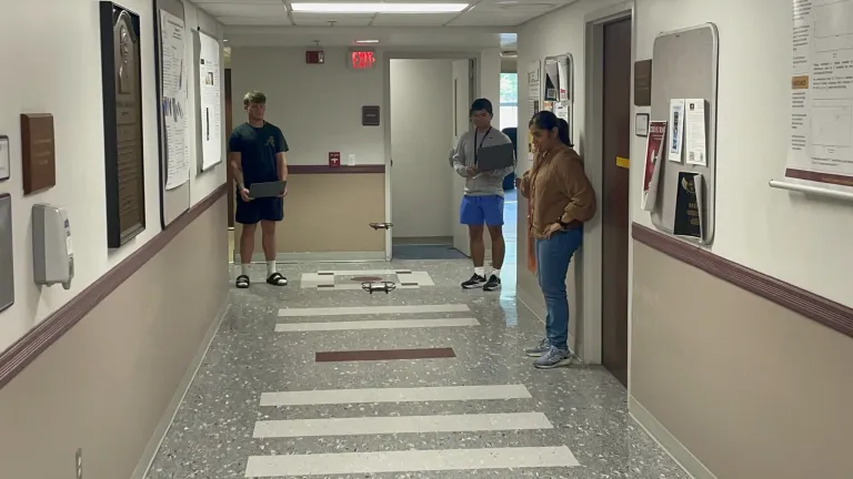 An instructor demonstrates a small drone to students seated around a classroom table with laptops.