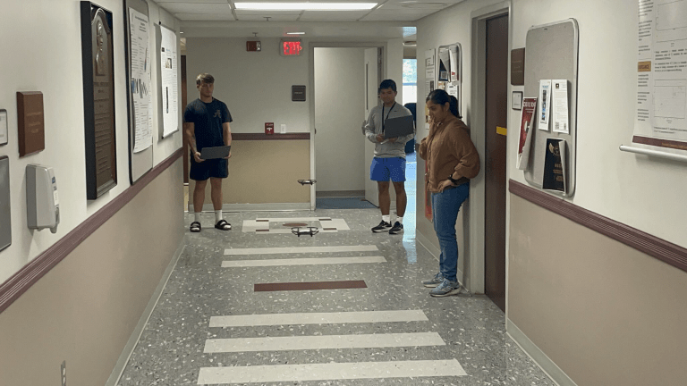 An instructor demonstrates a small drone to students seated around a classroom table with laptops.