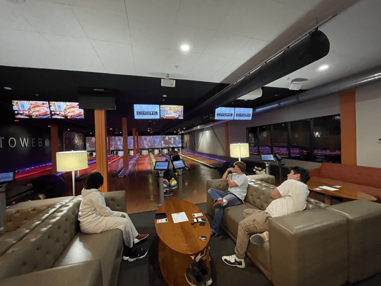 Students sit on couches watching bowling lanes inside a bowling alley.