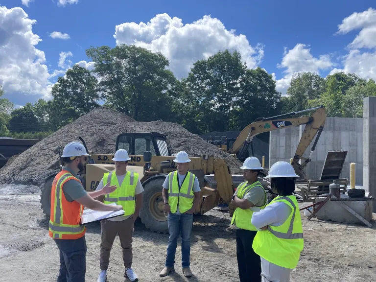 Students in hard hats and safety vests receive instruction at a construction site with heavy equipment.