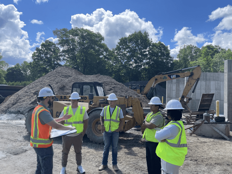 Students in hard hats and safety vests receive instruction at a construction site with heavy equipment.
