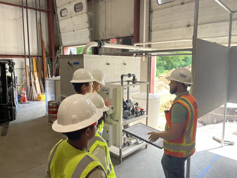Students in hard hats and safety vests listen to an instructor beside industrial equipment in a workshop.
