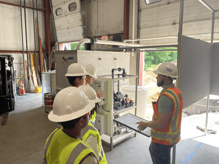 Students in hard hats and safety vests listen to an instructor beside industrial equipment in a workshop.