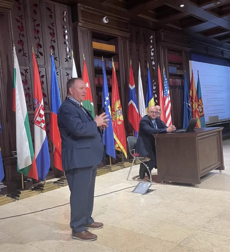 A speaker addresses an audience beside seated panelists with national flags displayed across the stage.