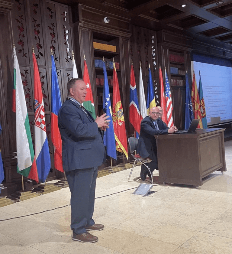 A speaker addresses an audience beside seated panelists with national flags displayed across the stage.