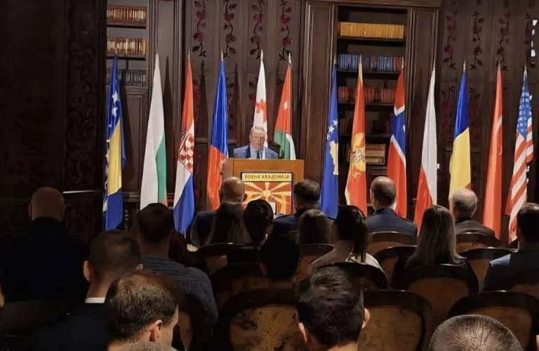 A speaker stands at a podium before an audience with national flags displayed behind the stage.
