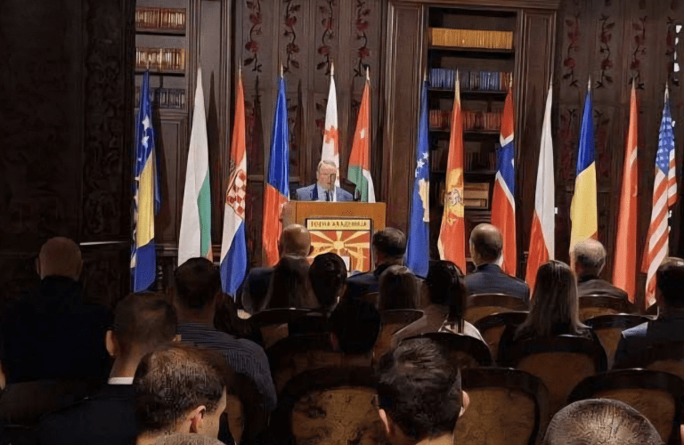 A speaker stands at a podium before an audience with national flags displayed behind the stage.