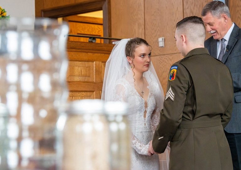 A bride and a groom in uniform hold hands during a wedding ceremony indoors at the Norwich Chapel.