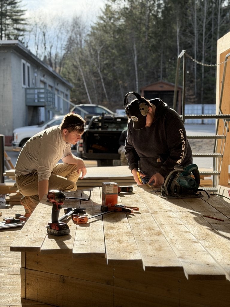 Two people measure and mark wooden boards on a workbench with tools nearby.