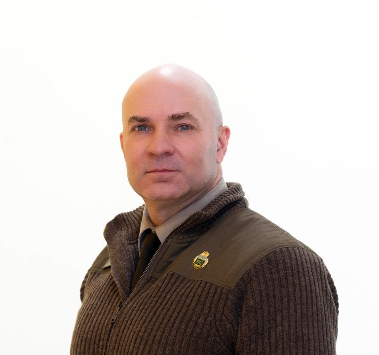 Person in a dark zip-up sweater and tie faces the camera against a white background.