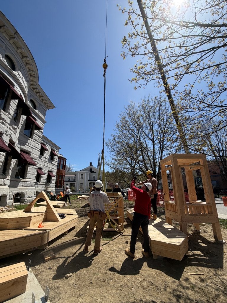 Workers guide a crane lifting a wooden play structure into place beside a building.