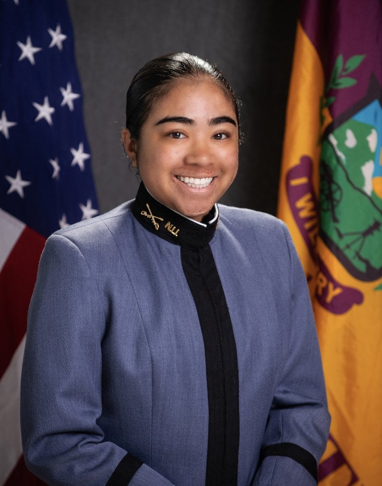 Cadet in dress uniform poses for a portrait between U.S. and Norwich University flags.