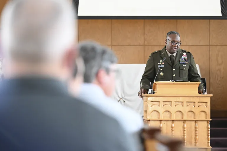 Uniformed officer speaks at a podium while audience members listen indoors.