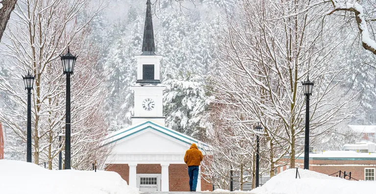 A person walks along a snow-covered path toward a chapel with a clock tower.