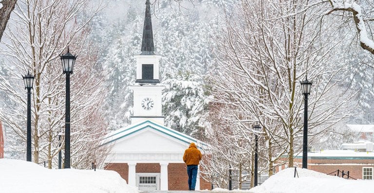 A person walks along a snow-covered path toward a chapel with a clock tower.